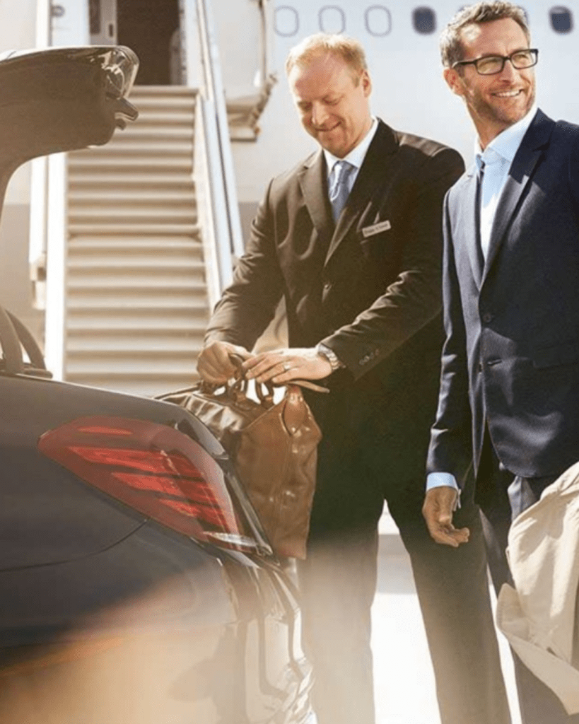 A chauffeur assisting a passenger with their luggage at an airport, with a luxury car in the foreground.