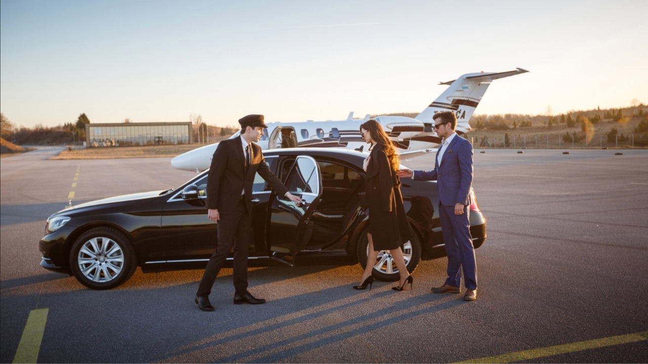 A chauffeur in a black suit is assisting a woman in an elegant black dress as she enters a luxury black sedan, while a man in a blue suit stands nearby. A private jet is visible in the background, indicating high-end travel.
