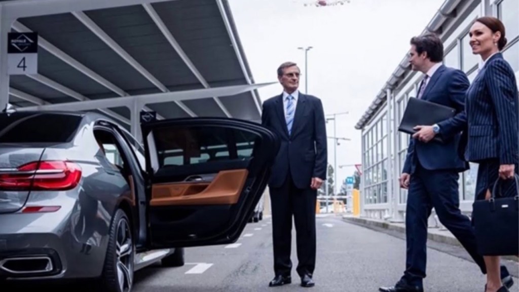 A professional chauffeur stands beside a luxury sedan with its door open, as two elegantly dressed individuals prepare to enter, likely at an airport or business venue.