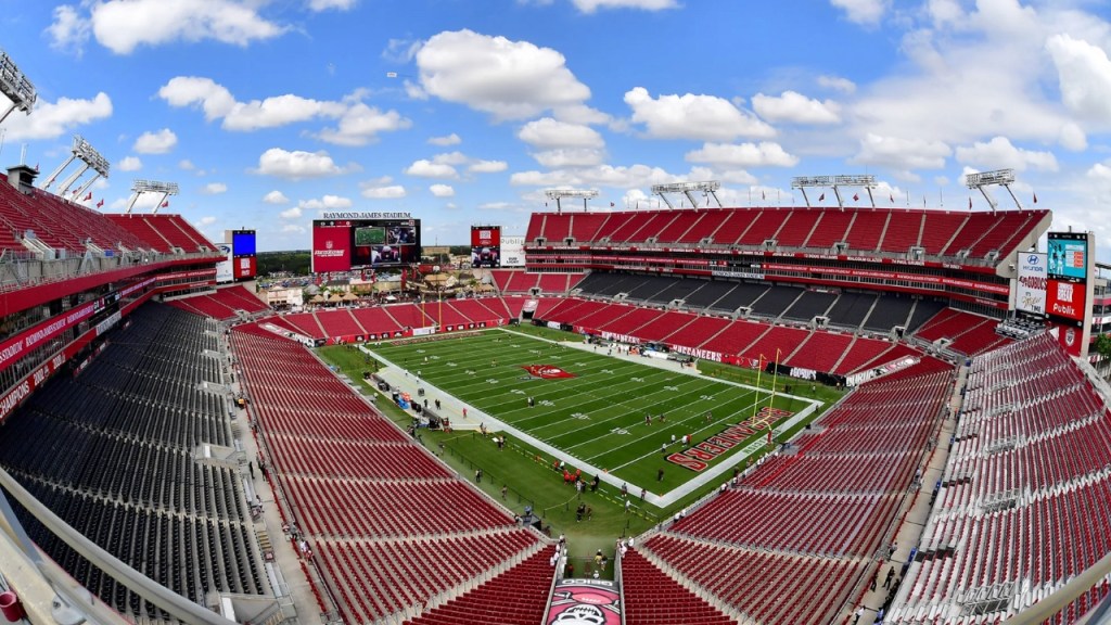 A panoramic view of Raymond James Stadium in Tampa Bay, showcasing the empty seating, a well-maintained football field, and a clear blue sky with scattered clouds.