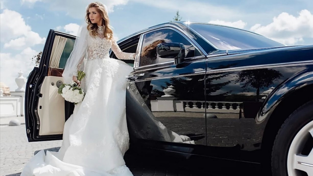 A bride in a stunning white wedding dress stepping out of a luxurious black limousine, holding a bouquet of white flowers. The scene is set against a bright blue sky.